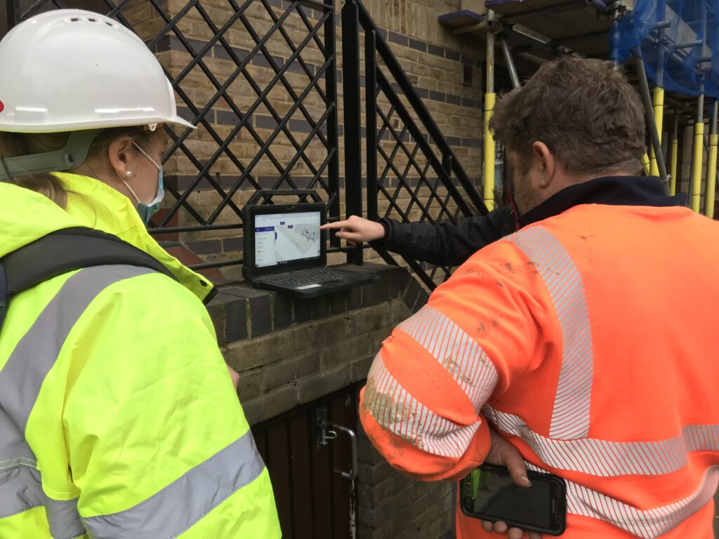 Two people in high-visibility jackets and a hard hat examine a digital site map on a rugged tablet outdoors, one pointing at the screen.
